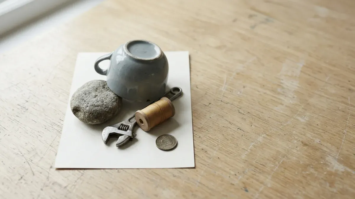A sheet of white paper on a scratched wooden desk, held down by five mismatched objects: an upturned ceramic cup, a stone, an adjustable wrench, a spool of thread and a coin.